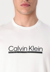 Camiseta blanca de algodón con un logo negro de "Calvin Klein" y una delgada línea horizontal debajo del texto. Diseño clásico de cuello redondo.