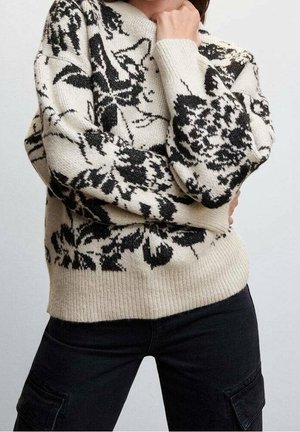 Person iført sorte cargobukser og en hvid strikket sweater med sort blomsterprint, poserer med armene krydsede og hånden nær ansigtet.