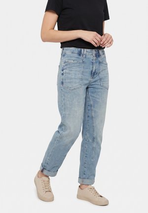 Personne portant un jean bleu clair avec revers, un t-shirt noir à manches courtes et des baskets beiges à lacets, debout, les mains légèrement croisées.