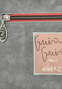 Graues Kunstledertäschchen mit einem roten Reißverschluss und einem silbernen Ziehband. Verziert mit einem beigen Etikett, auf dem handschriftlich "Guin Guin" und "anekke" steht.