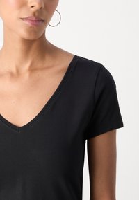 Black V-neck t-shirt in a lightweight, smooth fabric, with short sleeves and minimal stitching details. Accented by a large hoop earring.