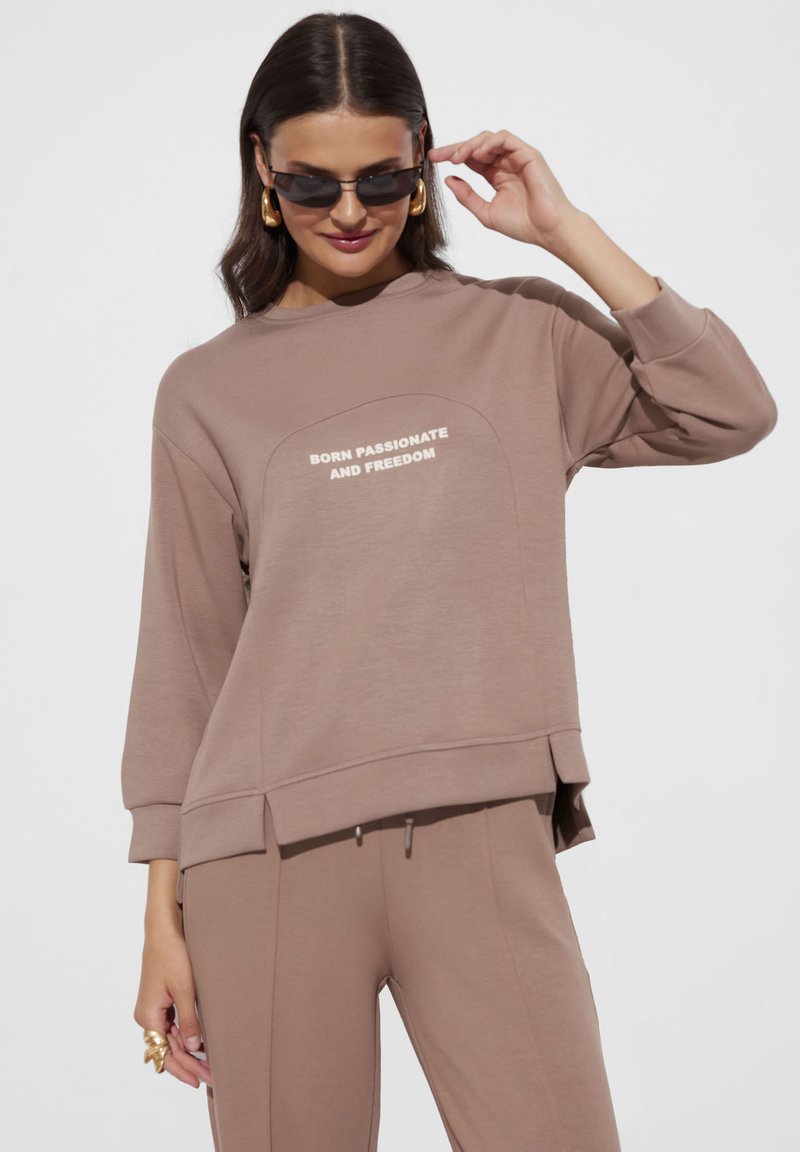 Brązowy, oversize'owy sweatshirt z białym napisem "URODZONY Z PASJĄ I WOLNOŚCIĄ", o luźnym kroju i bocznymi rozcięciami.