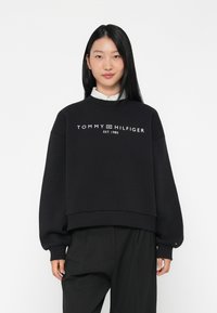 Juodas medvilninis džemperis su sutrumpintu dizainu. Priekyje yra balta logo tekstas „TOMMY HILFIGER EST. 1985“ ir gipiūriniai rankogaliai.