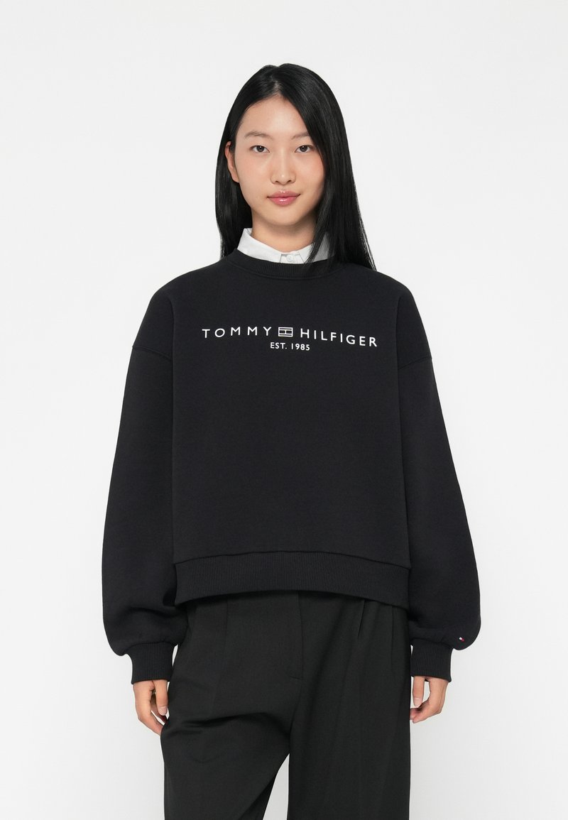 Juodas medvilninis džemperis su sutrumpintu dizainu. Priekyje yra balta logo tekstas „TOMMY HILFIGER EST. 1985“ ir gipiūriniai rankogaliai.