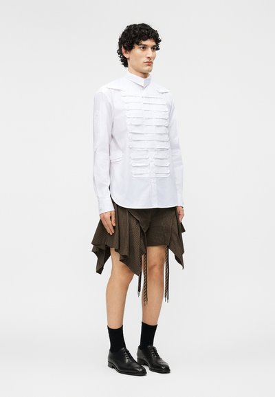 Młoda osoba stojąca, ubrana w białą koszulę z guzikami i poziomymi plisami oraz asymetryczną brązową spódnicę, czarne skarpetki i buty.