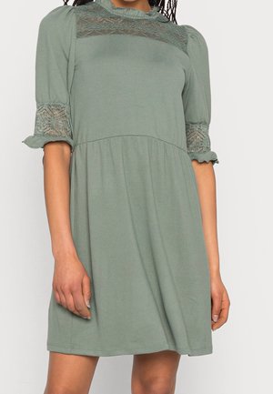 Femme portant une robe vert sauge avec des panneaux en dentelle sur la poitrine et les manches, avec des manches ballon mi-longues et une taille froncée.