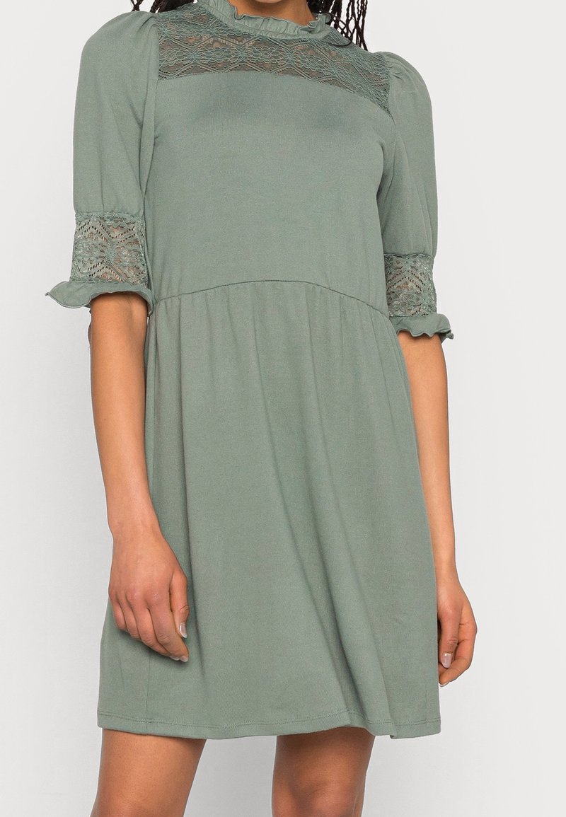 Femme portant une robe vert sauge avec des panneaux en dentelle sur la poitrine et les manches, avec des manches ballon mi-longues et une taille froncée.