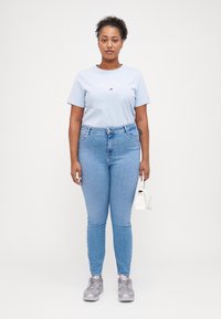 T-shirt bleu clair à manches courtes avec logo blanc, associé à un jean ajusté bleu clair et des baskets grises. Sac à main blanc tenu dans la main droite.