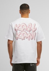 Balta kokvilnas T-kreklā ar īsām piedurknēm, uz muguras ir liels rozā burbuļu stila teksts "KAL" un "KANI". Minimalistisks dizains.