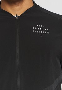 Zwarte sportjack met rits met "NIKE RUNNING DIVISION" in het wit gedrukt op de bovenste linkerborst, gedragen door een persoon.