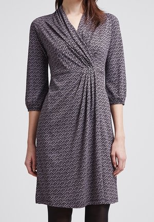 Femme portant une robe mi-longue à manches trois-quarts avec un motif géométrique violet et noir et un détail froncé à la taille.