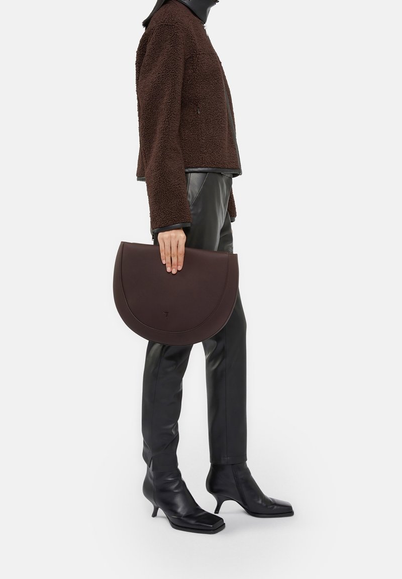 Personne portant une veste marron texturée, un pantalon en cuir noir, des bottes noires à talons, tenant un grand sac à main marron en forme de demi-lune à côté.