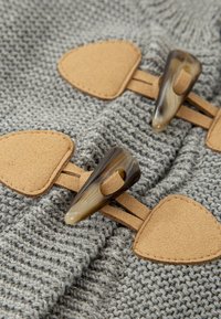 La tela de punto gris presenta dos cierres de gamuza color beige con puntas oscuras parecidas al cuerno, ofreciendo un contraste de texturas y un cierre seguro.