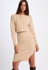 Abito maglione in beige, caratterizzato da una texture a trecce, orlo a coste, maniche lunghe e una fessura laterale, realizzato in materiale morbido e elasticizzato.