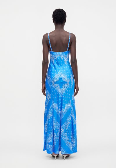 Femme portant une longue robe nuisette bleue à motifs floraux avec fines bretelles, debout de dos sur un fond blanc uni.