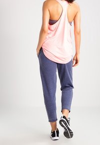 Top in rosa chiaro senza maniche con design a schiena incrociata, abbinato a joggers neri scuri. Scarpe sportive nere con suole bianche e dettagli testurizzati.