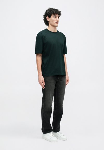 Sötétzöld, rövid ujjú póló, kerek nyakkivágással, sötét mosású, egyenes szárú farmernadrággal és fehér sportcipővel párosítva, semleges háttér előtt állva.