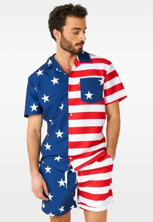 Mann trägt passende Shorts und ein kurzärmeliges Hemd mit amerikanischem Flaggen-Design; blau mit weißen Sternen auf der einen Seite, rot-weiße Streifen auf der anderen.