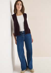 Ciemnofioletowy bezrękawnik z prążkowaną fakturą, z guzikami, noszony na białej bluzce, w połączeniu z niebieskimi spodniami o szerokich nogawkach i beżowymi butami.