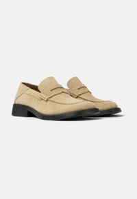 Beige suede loafers featuring a moccasin-style design, stitched accents, and a low black rubber sole. Smooth texture with rounded toe shape.