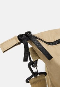 Beige olkalaukku, jossa on musta vetoketju vetotapeilla, rakenteellinen kangas ja kestävä klipsikiinnitys turvallista sulkemista varten.
