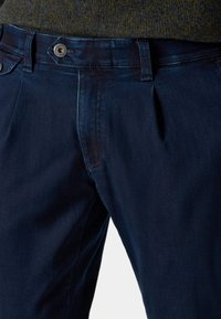 Dunkelblaue Jeanshose aus Denim mit flachem Vorderteil, seitlichen Taschen und einem Metallknopfverschluss am Bund. Glatte Textur.