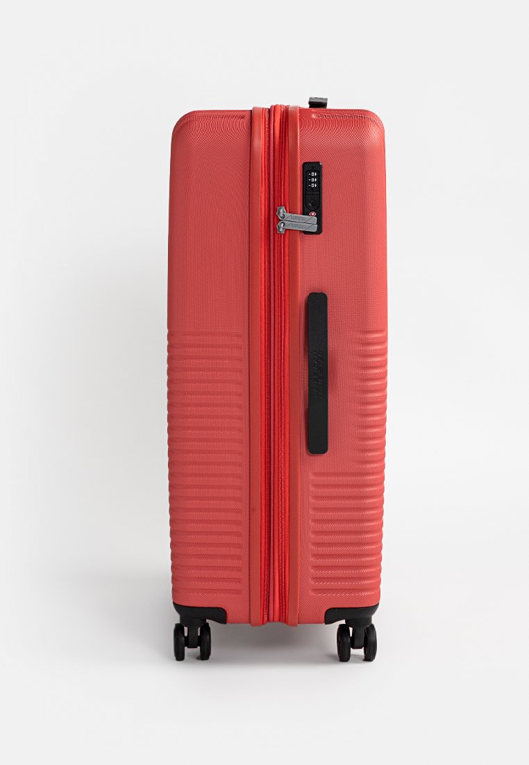 Valise rigide rouge se tenant debout avec quatre roues noires, une poignée latérale noire et un cadenas à combinaison intégré sur la fermeture éclair.