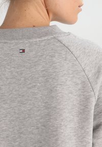 Světle šedá mikina s kulatým výstřihem a raglánovými rukávy, texturovaná tkanina, s malým logem v červené, bílé a modré barvě na zadní straně.