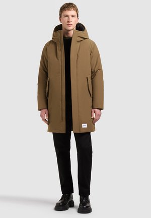 Young male model wearing a brown hooded winter coat, black pants, and black boots, standing on a light gray background.