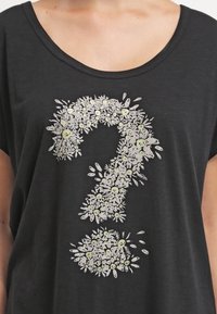 Černé tričko s krátkým rukávem, na kterém je velký bílý design květinového otazníku, obsahující usmívající se tváře a zelené akcenty. Měkká, volná textura.