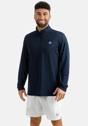 Uomo sorridente che indossa una maglia blu navy a maniche lunghe con zip a quattro, e pantaloncini bianchi con piccoli loghi sul petto e sulla gamba, in piedi davanti a uno sfondo bianco.