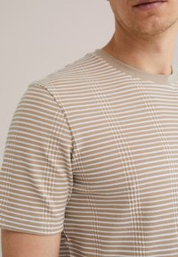 Béžové tričko s krátkým rukávem vyrobené z texturované tkaniny, které má tenké bílé pruhy. Hladký výstřih s jemným geometrickým vzorem.