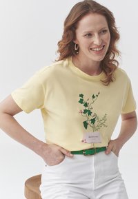 Camiseta de algodón amarilla con estampado de enredaderas verdes y etiqueta de texto 'ENREDADERAS HERMOSAS'. Combinada con pantalones blancos y un cinturón verde. Taburete de madera en el fondo.