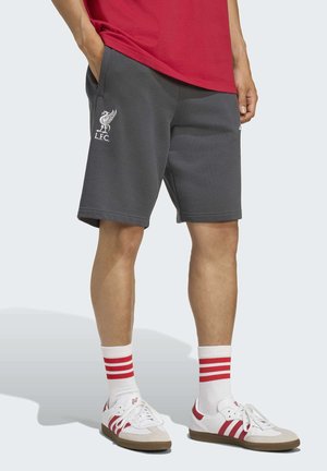 Homme portant un short Liverpool FC gris foncé, des chaussettes blanches avec des rayures rouges, des baskets blanches avec des détails rouges, et un t-shirt rouge.