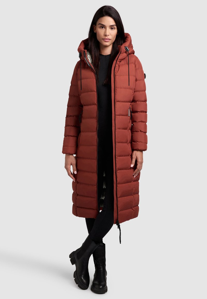 Brown puffer coat with a hood, vertical quilted design, zip closure, and side pockets. Worn over a black outfit and paired with black ankle boots.