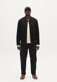 Črna jakna z dvema sprednjima žepoma, pod njo bela srajca, temni hlače in rjavi čevlji. Material se zdi teksturiran s trdim vzorcem.