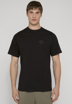 Mand iført en simpel sort t-shirt med korte ærmer og et lille logo på brystet, stående mod en lysegrå baggrund.