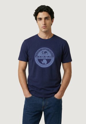 Giovane uomo con capelli ricci scuri che indossa una maglietta grafica blu navy e jeans blu, in piedi con le mani in tasca davanti a uno sfondo semplice.