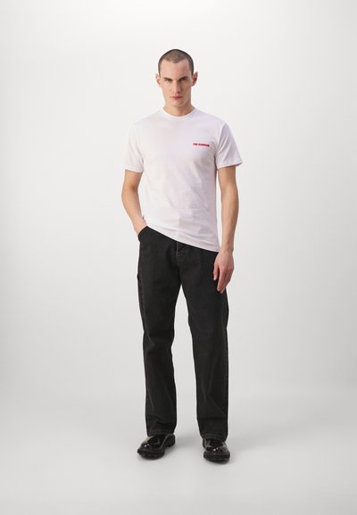Camiseta blanca de algodón con texto rojo "THE KOOPLES" en el lado izquierdo del pecho, combinada con pantalones negros y zapatos negros brillantes.