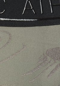 Grüner Stoff mit einer subtilen Textur, der an der Oberseite ein schwarzes elastisches Band aufweist. Das Band hat geprägte weiße Buchstaben. Ein dekoratives hellgraues Muster ist enthalten.