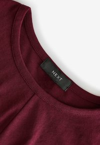 Burgundy puuvillapaita pyöreällä kaula-aukolla, jossa on sauma-aitoja ja musta etiketti, jossa on brändin "NEXT" ommeltu sisään.