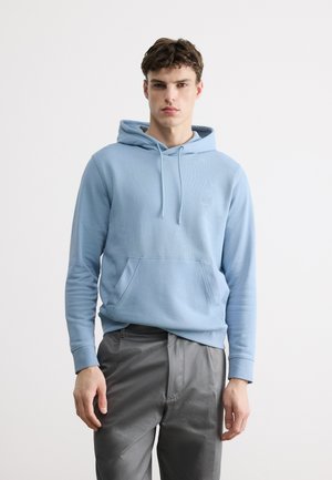 Młody mężczyzna ubrany w jasnoniebieską bluzę z kapturem i szare spodnie, stojący na tle białego, jednolitego tła.