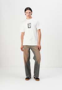 Biele tričko s krátkym rukávom a čiernym grafickým potlačou, doplnené vyblednutými svetlohnedými širokými džínsami a čiernymi teniskami s hnedými podrážkami.