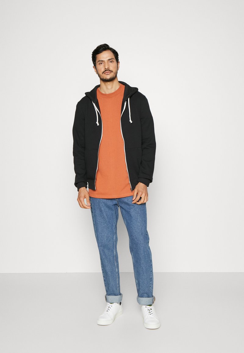 Zwarte zip-up hoodie met witte trekkoorden over een oranje T-shirt, gecombineerd met opgerolde blauwe jeans en witte sneakers.