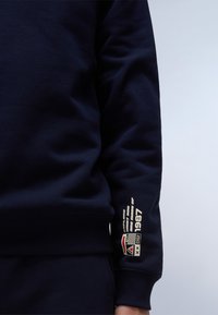 Marineblauwe sweatshirt met geribbelde manchetten. Heeft een creme kleurige graphic op de mouw met de tekst "1987" en patroonaccenten. Zachte textuur.