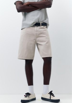 Homme debout, les bras croisés, portant un short beige, une chemise rayée, des chaussettes blanches et des baskets blanches avec des semelles et des lacets noirs.