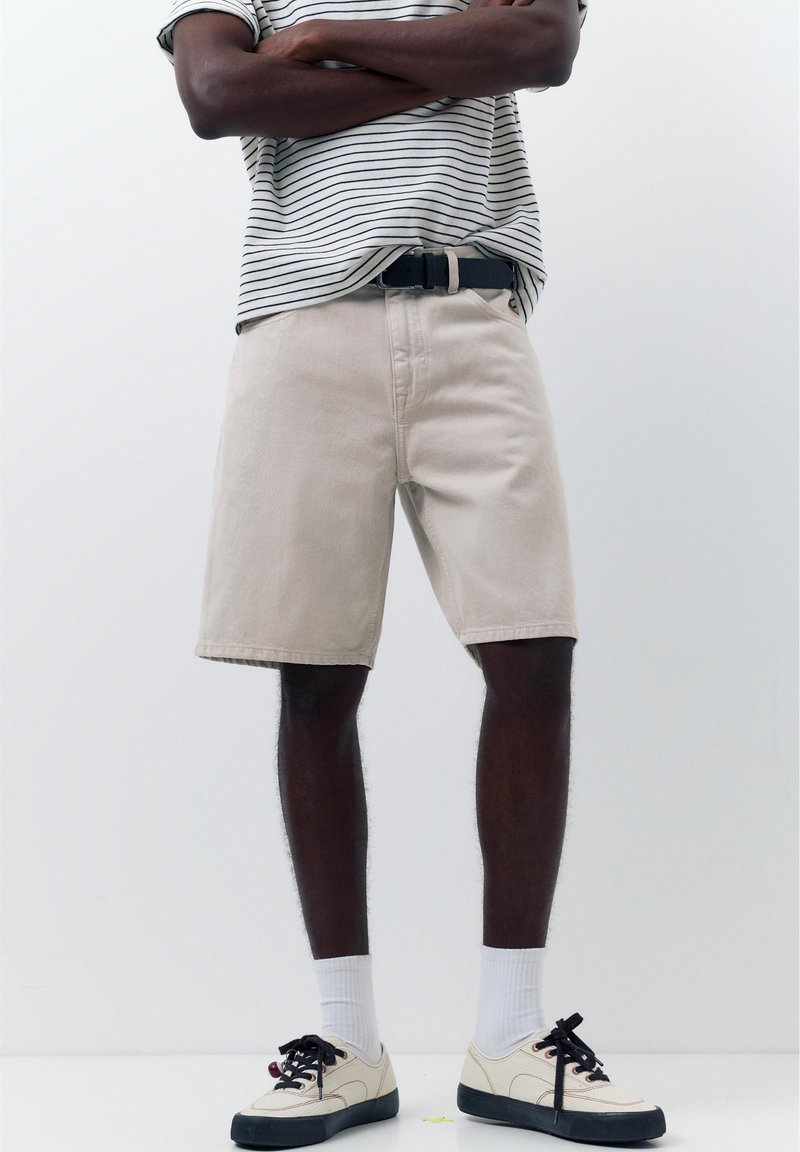 Homme debout, les bras croisés, portant un short beige, une chemise rayée, des chaussettes blanches et des baskets blanches avec des semelles et des lacets noirs.