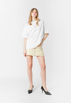 Camiseta blanca de gran tamaño con cuello redondo, combinada con una falda corta amarilla de capas que presenta un zipper frontal y múltiples secciones horizontales. Zapatos de tacón negros.