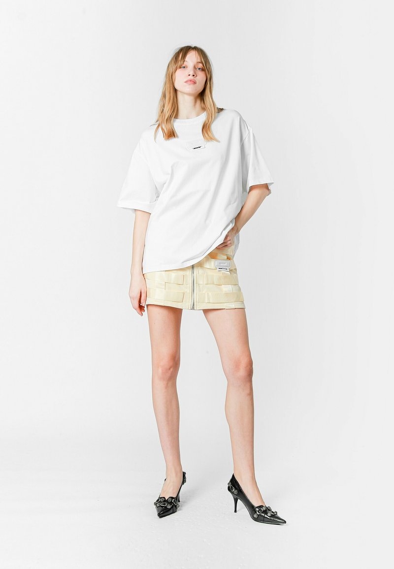Camiseta blanca de gran tamaño con cuello redondo, combinada con una falda corta amarilla de capas que presenta un zipper frontal y múltiples secciones horizontales. Zapatos de tacón negros.