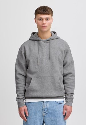 Ung mand med kort brunt hår iført en ensfarvet grå hættetrøje og lyseblå jeans, stående foran en hvid baggrund.